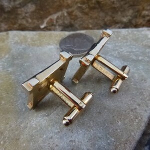 May include: A pair of gold-tone cufflinks with a square design. The cufflinks have a textured surface and are marked "D&M" on the back.