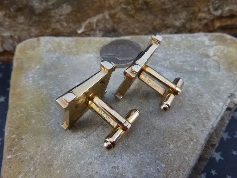 May include: A pair of gold-tone cufflinks with a square design. The cufflinks have a textured surface and are marked "D&M" on the back.