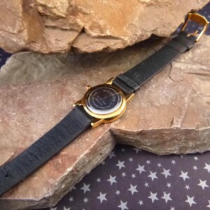 Op de afbeelding: Een goudkleurig Movado horloge met een zwarte leren band. Het horloge ligt op een getextureerde, lichtbruine rots. De wijzerplaat is zichtbaar en de band is vastgemaakt met een gouden gesp. De achtergrond is een donkerblauwe ondergrond met witte sterren.