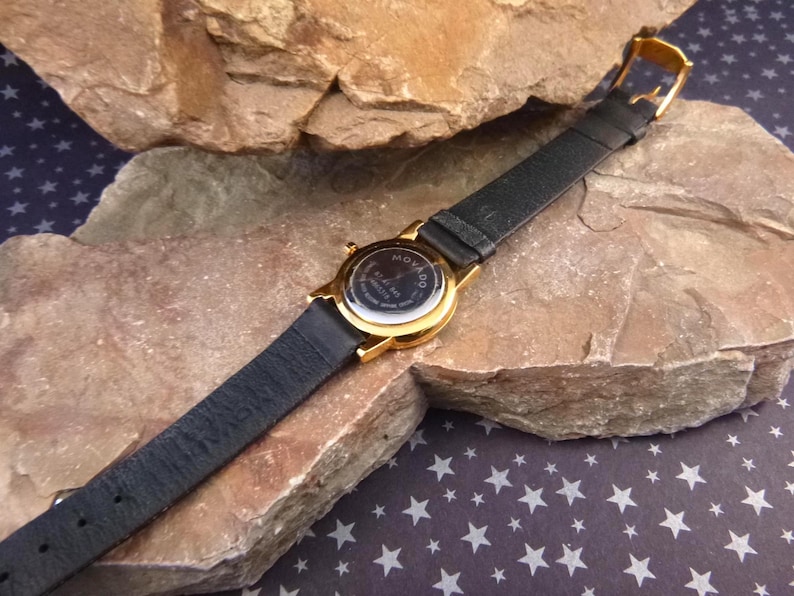 Op de afbeelding: Een goudkleurig Movado horloge met een zwarte leren band. Het horloge ligt op een getextureerde, lichtbruine rots. De wijzerplaat is zichtbaar en de band is vastgemaakt met een gouden gesp. De achtergrond is een donkerblauwe ondergrond met witte sterren.
