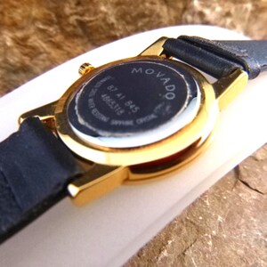 Op de afbeelding: Close-up van een goudkleurig Movado horloge met een zwarte leren band. De wijzerplaat van het horloge is zwart met witte tekst, inclusief de merknaam en modeldetails. Het horloge staat op een neutrale achtergrond.