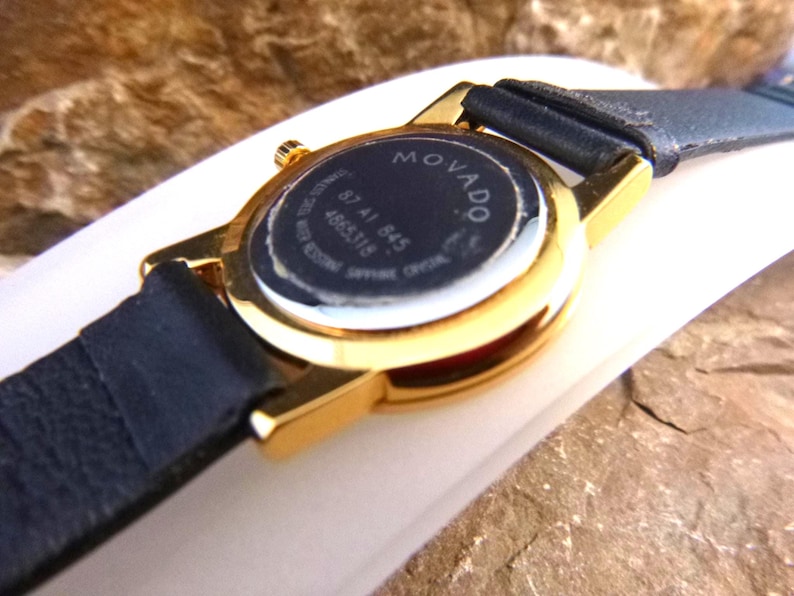Op de afbeelding: Close-up van een goudkleurig Movado horloge met een zwarte leren band. De wijzerplaat van het horloge is zwart met witte tekst, inclusief de merknaam en modeldetails. Het horloge staat op een neutrale achtergrond.