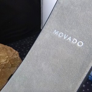 Op de afbeelding: Een close-up van een grijze, getextureerde etui met de merknaam "MOVADO" in het wit geborduurd. De etui is open en onthult een glimp van een horloge of sieraad. De achtergrond is donker.
