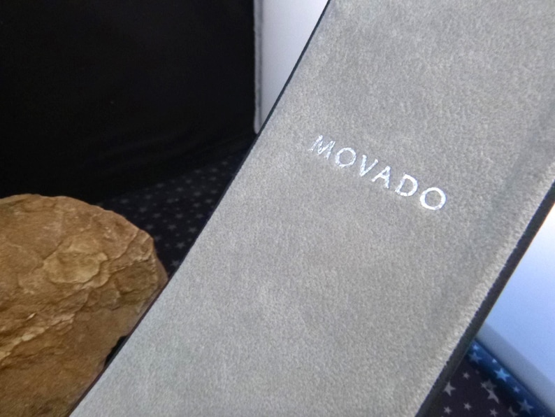 Op de afbeelding: Een close-up van een grijze, getextureerde etui met de merknaam "MOVADO" in het wit geborduurd. De etui is open en onthult een glimp van een horloge of sieraad. De achtergrond is donker.