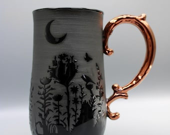 May include: A dark gray ceramic mug with a copper-colored handle. The mug features a black silhouette design of a moon, flowers, and a butterfly. The handle has an ornate, branch-like design.
