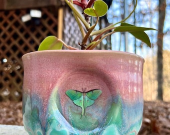 May include: A green and pink ceramic planter with a green luna moth design. The planter has a drip glaze and a matching saucer.