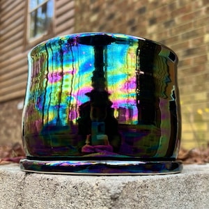 May include: A ceramic planter with a glossy, iridescent finish. The planter is a dark color, reflecting a spectrum of colors including blue, green, and purple. It sits on a matching saucer, suitable for indoor or outdoor use.