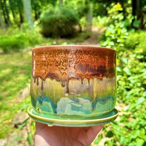 May include: A handcrafted ceramic planter with a rich, glazed finish. The top is a warm brown, transitioning to green and turquoise hues towards the base. The planter sits on a matching saucer, perfect for indoor or outdoor use. A unique piece for home decor.