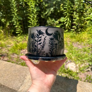 May include: A ceramic planter with a black floral design and a crescent moon. The planter is gray with a black rim and sits on a matching saucer. The design features silhouettes of flowers and foliage.