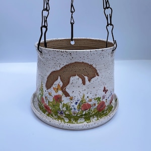 May include: A hanging ceramic planter with a speckled white finish. It features a brown fox illustration and a floral design with red poppies and butterflies. The planter is suspended by black chains.
