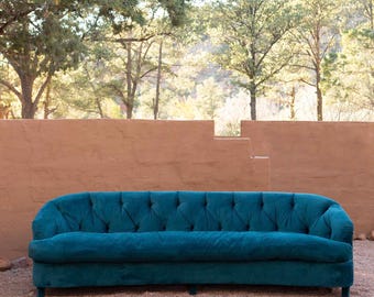 May include: A teal velvet sofa with a curved back and button tufting. The sofa is placed outdoors on a gravel surface, with a terracotta-colored wall and trees in the background.