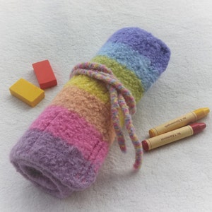 Op de afbeelding: Een opgerolde, regenboogkleurige etui met een bijpassende strik. De etui is gemaakt van een zacht, getextureerd materiaal en is versierd met banden in paars, roze, oranje, geel en groen. Twee kleurpotloden en twee rechthoekige gummen liggen ernaast.