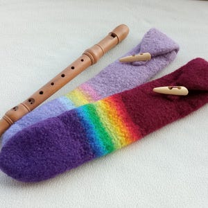Könnte beinhalten: Eine Holzblockflöte mit zwei farbigen Filzetuis. Die Etuis sind lila, regenbogenfarben und weinrot, jeweils mit einem Knebelverschluss aus Holz. Die Blockflöte ist hellbraun und hat mehrere Fingerlöcher.
