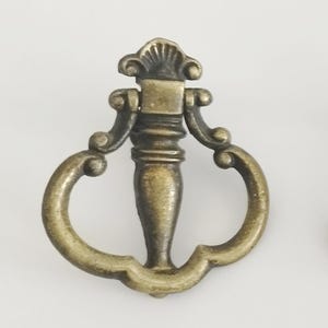 Peut inclure: Trois poignées de tiroir en laiton antique avec un design décoratif. Chaque poignée présente un poteau central avec une poignée incurvée et des détails ornés. Les poignées sont disposées horizontalement sur un fond blanc.