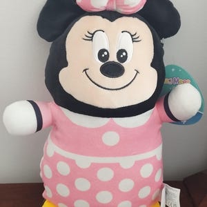 Peut inclure: Une peluche Minnie Mouse. La poup&eacute;e a une t&ecirc;te noire avec de grandes oreilles, un n&oelig;ud rose &agrave; pois blancs et un visage souriant. Elle porte une robe rose &agrave; pois blancs et des chaussures jaunes.