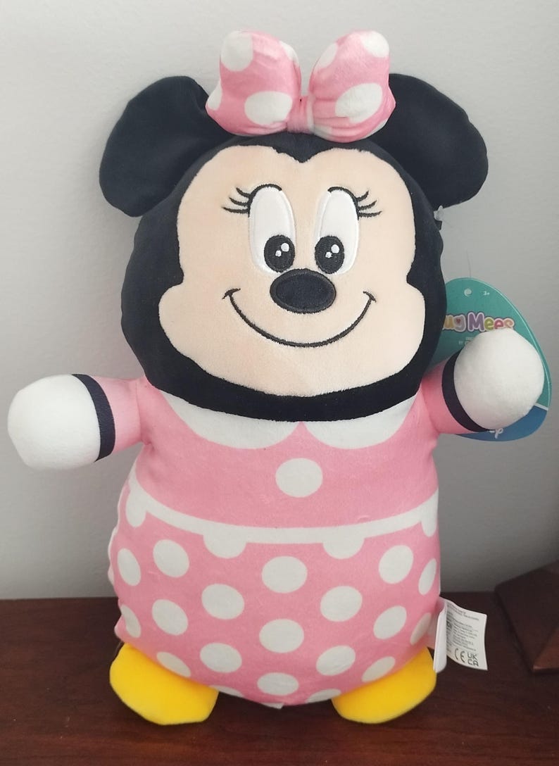 Peut inclure: Une peluche Minnie Mouse. La poup&eacute;e a une t&ecirc;te noire avec de grandes oreilles, un n&oelig;ud rose &agrave; pois blancs et un visage souriant. Elle porte une robe rose &agrave; pois blancs et des chaussures jaunes.