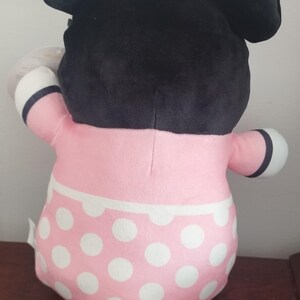 Peut inclure: Une poup&eacute;e en peluche de Minnie Mouse, vue de dos, avec une t&ecirc;te noire, de grandes oreilles et un n&oelig;ud rose &agrave; pois blancs. La poup&eacute;e porte une robe rose &agrave; pois blancs et des d&eacute;tails noirs et blancs.