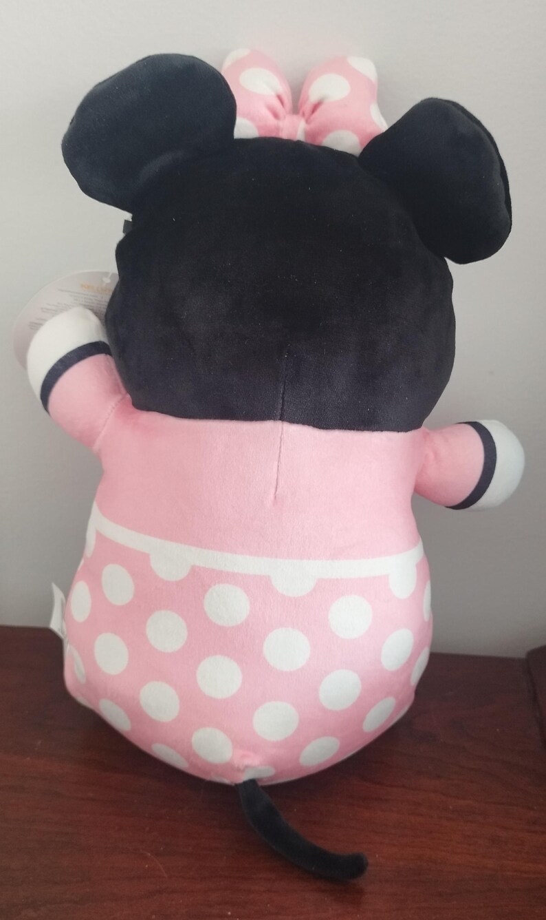 Peut inclure: Une poup&eacute;e en peluche de Minnie Mouse, vue de dos, avec une t&ecirc;te noire, de grandes oreilles et un n&oelig;ud rose &agrave; pois blancs. La poup&eacute;e porte une robe rose &agrave; pois blancs et des d&eacute;tails noirs et blancs.