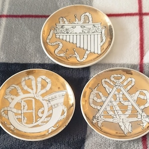 May include: Three small, round, decorative plates with gold rims and centers. Each plate features a different white and gold musical instrument design. The plates are arranged on a patterned surface.