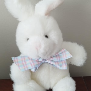 May include: A fluffy white bunny toy with a pastel plaid bow. The bunny has long ears, black eyes, and a pink nose. The soft toy is designed for children.