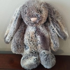 May include: A soft toy bunny rabbit in shades of grey, brown and white. The bunny has long, floppy ears and a pink nose. The plush toy has a textured fur appearance and is sitting upright. A cuddly stuffed animal.