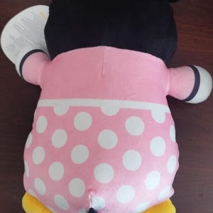 Peut inclure: Une poup&eacute;e en peluche Minnie Mouse, vue de dos, avec une t&ecirc;te noire avec de grandes oreilles, une robe rose &agrave; pois blancs et des pieds jaunes. La poup&eacute;e a une queue noire et des gants blancs.