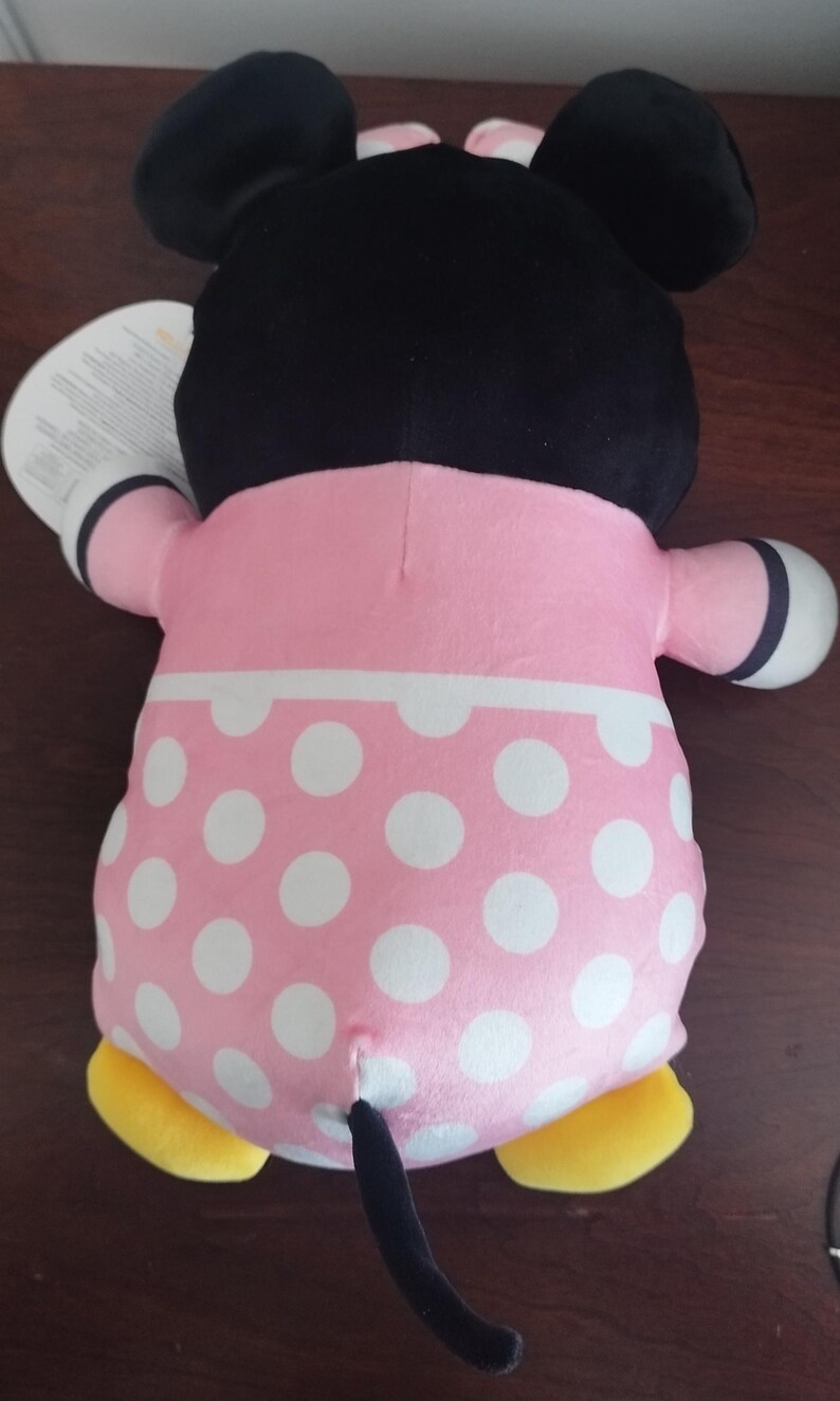 Peut inclure: Une poup&eacute;e en peluche Minnie Mouse, vue de dos, avec une t&ecirc;te noire avec de grandes oreilles, une robe rose &agrave; pois blancs et des pieds jaunes. La poup&eacute;e a une queue noire et des gants blancs.