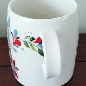 May include: White ceramic mug with a curved handle and a floral design. The mug features a colorful pattern of red, blue, and green leaves and flowers. The mug has a smooth, glossy finish and is suitable for hot or cold beverages.