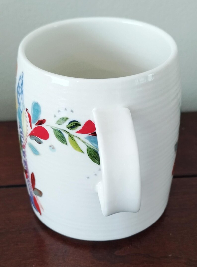 May include: White ceramic mug with a curved handle and a floral design. The mug features a colorful pattern of red, blue, and green leaves and flowers. The mug has a smooth, glossy finish and is suitable for hot or cold beverages.