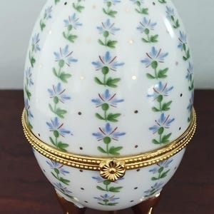 May include: A white porcelain egg-shaped trinket box with a hinged lid and gold-coloured metal accents. The egg is decorated with blue floral patterns and gold dots. It stands on three gold-coloured feet.
