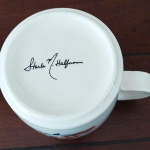 May include: White ceramic mug with a handle, featuring a floral design in red and blue. The bottom of the mug has a signature in black ink. The mug is set against a dark wooden surface.