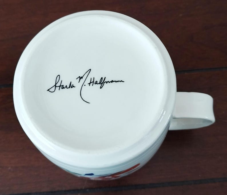 May include: White ceramic mug with a handle, featuring a floral design in red and blue. The bottom of the mug has a signature in black ink. The mug is set against a dark wooden surface.