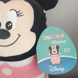 Peut inclure: Peluche Minnie Mouse avec une robe rose &agrave; pois blancs et un n&oelig;ud. Elle a des oreilles noires et un visage souriant. L'&eacute;tiquette indique "HugMees" et "Minnie Mouse" avec le logo Disney.