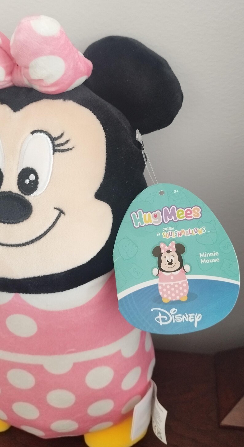 Peut inclure: Peluche Minnie Mouse avec une robe rose &agrave; pois blancs et un n&oelig;ud. Elle a des oreilles noires et un visage souriant. L'&eacute;tiquette indique "HugMees" et "Minnie Mouse" avec le logo Disney.
