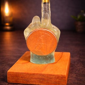 May include: A unique bottle stopper shaped like a hand giving the middle finger. The clear resin hand holds a copper-colored coin and is mounted on a square wooden base. The top of the finger has a gold-colored cap.