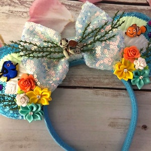 May include: Blue and green sequined Minnie Mouse ears with a bow, featuring a Finding Nemo theme. The ears are decorated with a variety of flowers, including orange, yellow, and white. The bow is made of iridescent sequins and has a small turtle charm. The ears also feature a small Dory character and a small Nemo character.