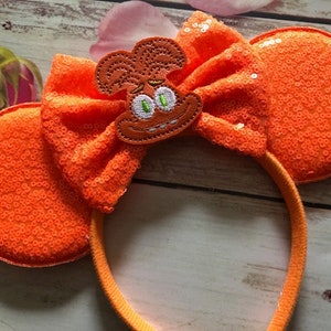 May include: Orange sequined Minnie Mouse ears with a brown felt character appliqué. The character has a surprised expression with green eyes and a brown mouth.