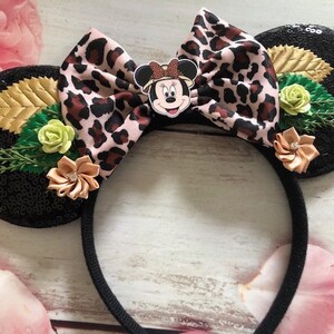 May include: Black mouse ears with a leopard print bow, green leaves, and a brown flower. The bow has a small Minnie Mouse character in the center.