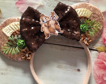Op de afbeelding: Een paar bruine en roosgouden muizenoren met een bruine strik en een chipmunk- en dale-personage-applicatie. De oren zijn versierd met bladeren en bloemen.