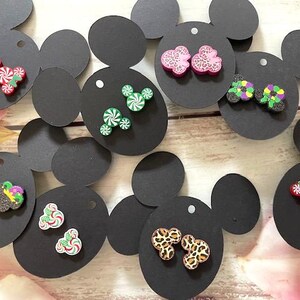 May include: Assortment of colorful, whimsical earrings displayed on black Mickey Mouse-shaped cards. The earrings feature various designs, including candy canes, Mickey Mouse heads, and animal prints.