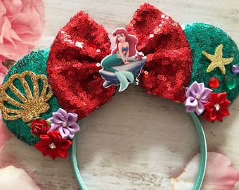 May include: Green and red sequined Minnie Mouse ears with a mermaid character, seashells, and flowers.