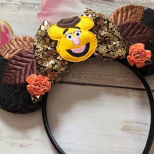 May include: A black headband with two large, sequined ears. The ears are decorated with brown leaves, orange roses, and a yellow character with a brown hat. A gold sequined bow sits between the ears.