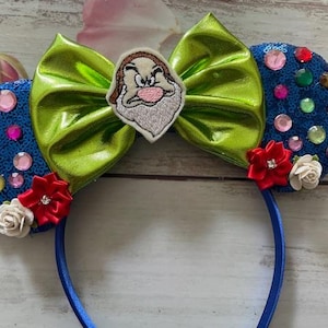 May include: A blue headband with sequined ears adorned with colorful gems and small red and white flowers. A large green bow sits in the center, with an embroidered image of Grumpy from Snow White.