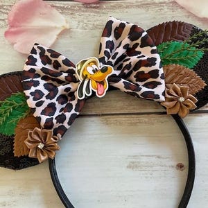 May include: Black and brown mouse ears with a leopard print bow, brown leaves, and a Goofy character appliqué.