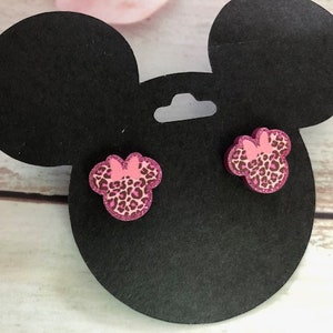 May include: A pair of pink and black glitter Minnie Mouse earrings. The earrings are shaped like Minnie Mouse's ears and have a pink bow on top.