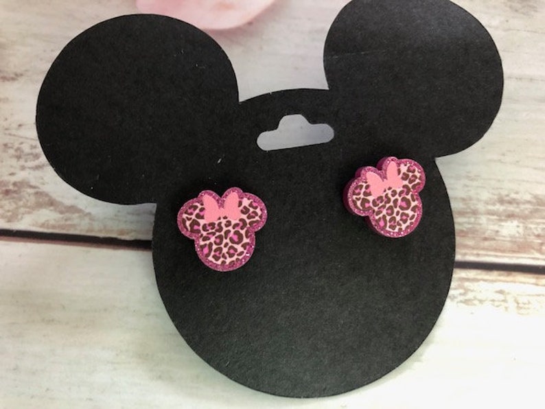 May include: A pair of pink and black glitter Minnie Mouse earrings. The earrings are shaped like Minnie Mouse's ears and have a pink bow on top.