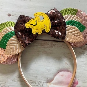 May include: A pair of brown and pink sequined Minnie Mouse ears with a yellow felt lion applique. The ears are decorated with green and beige felt leaves.