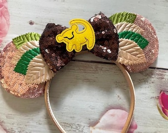 Op de afbeelding: Een paar bruine en roze pailletten Minnie Mouse oren met een gele vilt leeuwen applique. De oren zijn versierd met groene en beige vilt bladeren.
