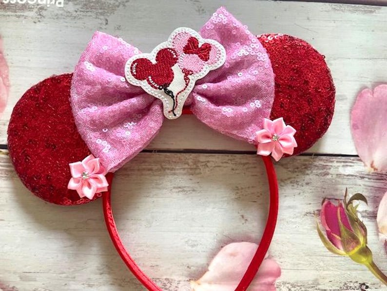 Puede incluir: Una diadema roja con orejas de lentejuelas y un lazo rosa de lentejuelas. El lazo presenta un dise&ntilde;o bordado de globos de Mickey Mouse. Acentos de flores rosas a cada lado del lazo.