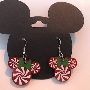 May include: A pair of red and white peppermint candy cane themed dangle earrings with green bows. The earrings are shaped like Mickey Mouse ears.